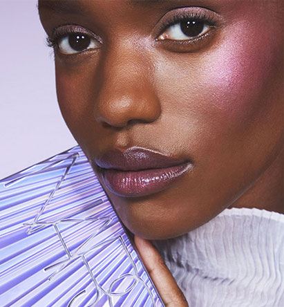 A close-up of a woman with shimmering pink makeup on her eyelids, cheeks, and lips. She is holding a reflective, lavender-colored object and wearing a white, ribbed turtleneck. The background is a soft lavender shade.
