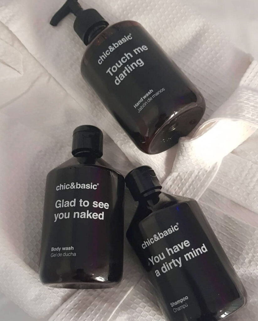 Three black bottles of chic&basic toiletries with playful white text labels, placed on a white textured towel. The bottles are labeled: "Touch me darling," "Glad to see you naked," and "You have a dirty mind.