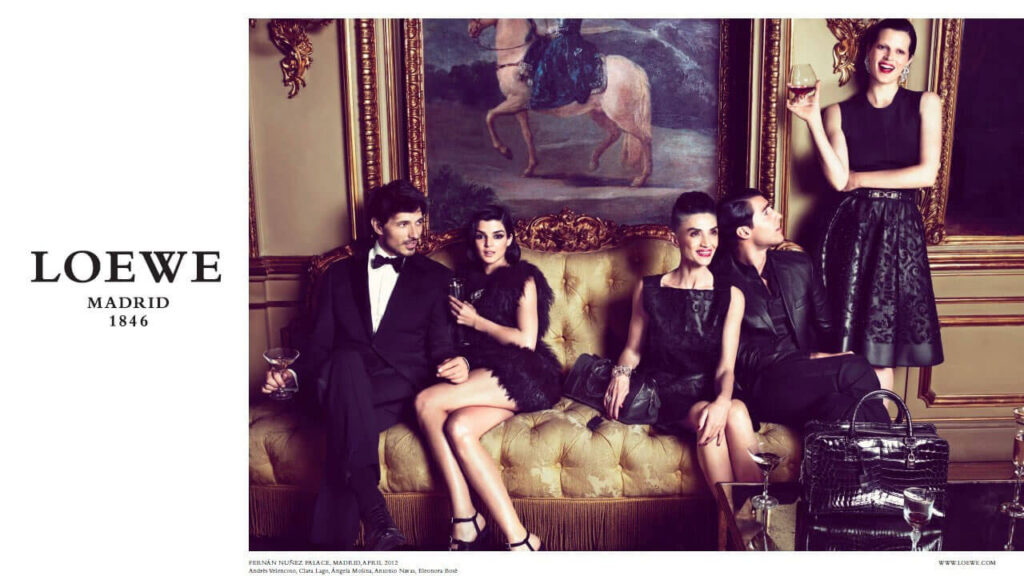 A fashionably dressed group of five people sit and stand around an ornate gold sofa in an elegant room, holding drinks. The scene exudes luxury and sophistication. "Loewe Madrid 1846" is written on the left.
