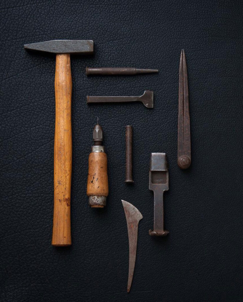 A set of vintage leatherworking tools, including a wooden-handled hammer, awl, chisels, and other specialized metal tools, arranged neatly on a textured black surface.